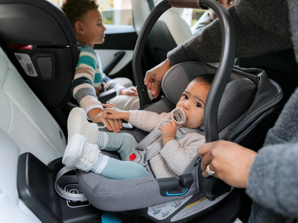 Toddler and baby hold hands while securely strapped into the Alta V2 and Aria, respectively. 