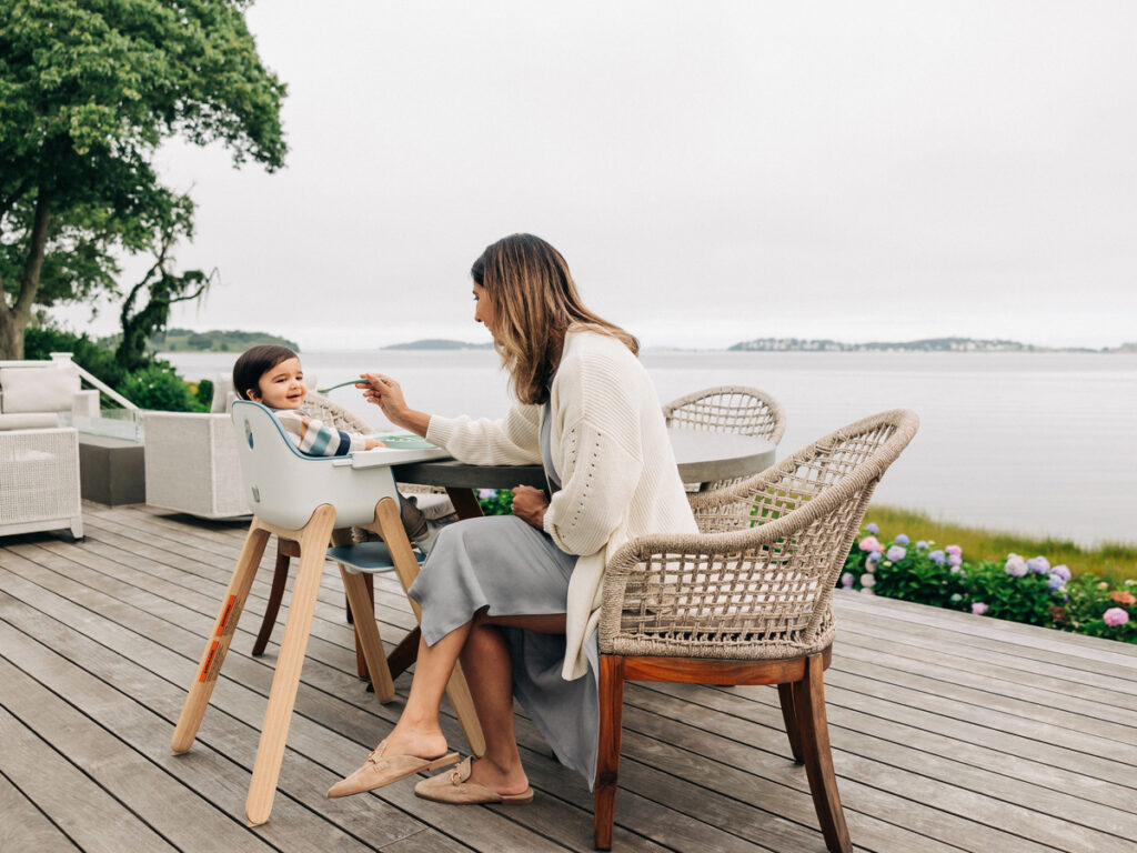 A mom feeds her child pulled right up to the table in the Ciro High Chair, both of them enjoying mealtime together on a beautiful day