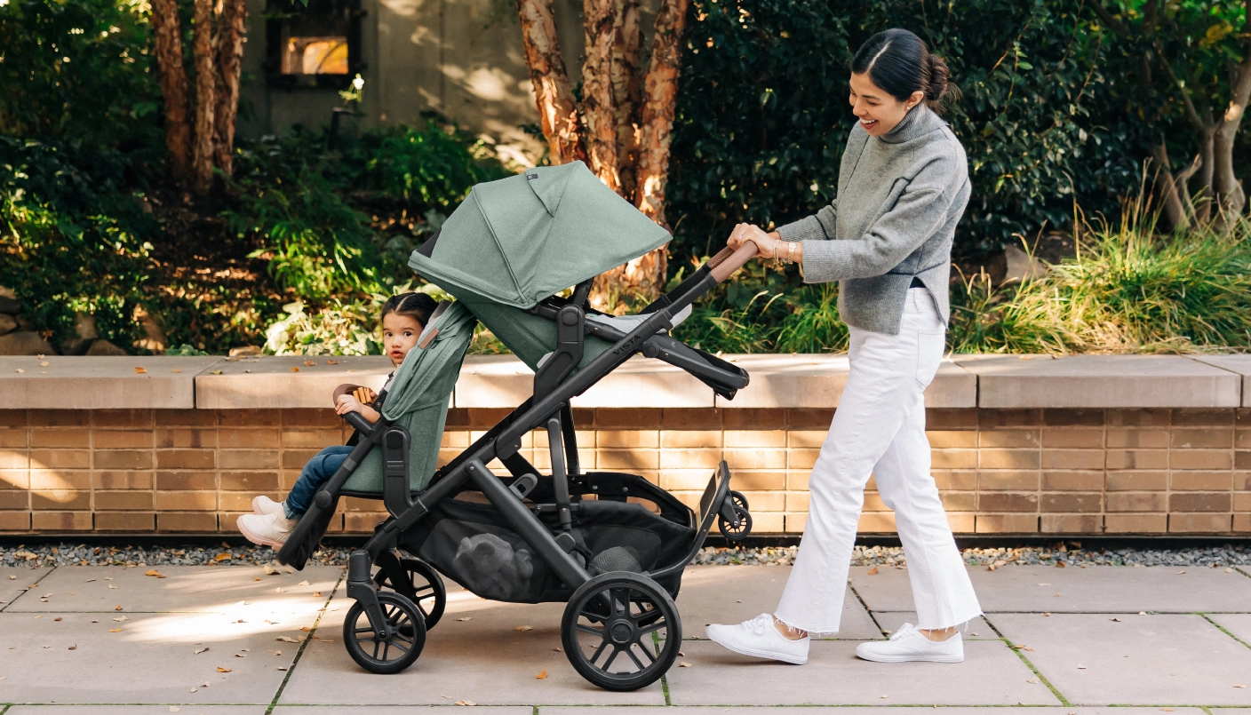 Parent smiling while pushing Vista Stroller with forward facing RumbleSeat V2+ and parent facing Toddler Seat all in Gwen fashion