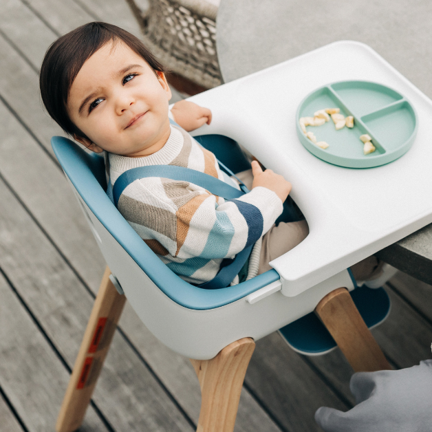 A baby smiles and looks to the camera while secured in the Ciro High Chair, which features a seam-free seat and removable, food-friendly harness for easy and smooth wipe down and cleaning