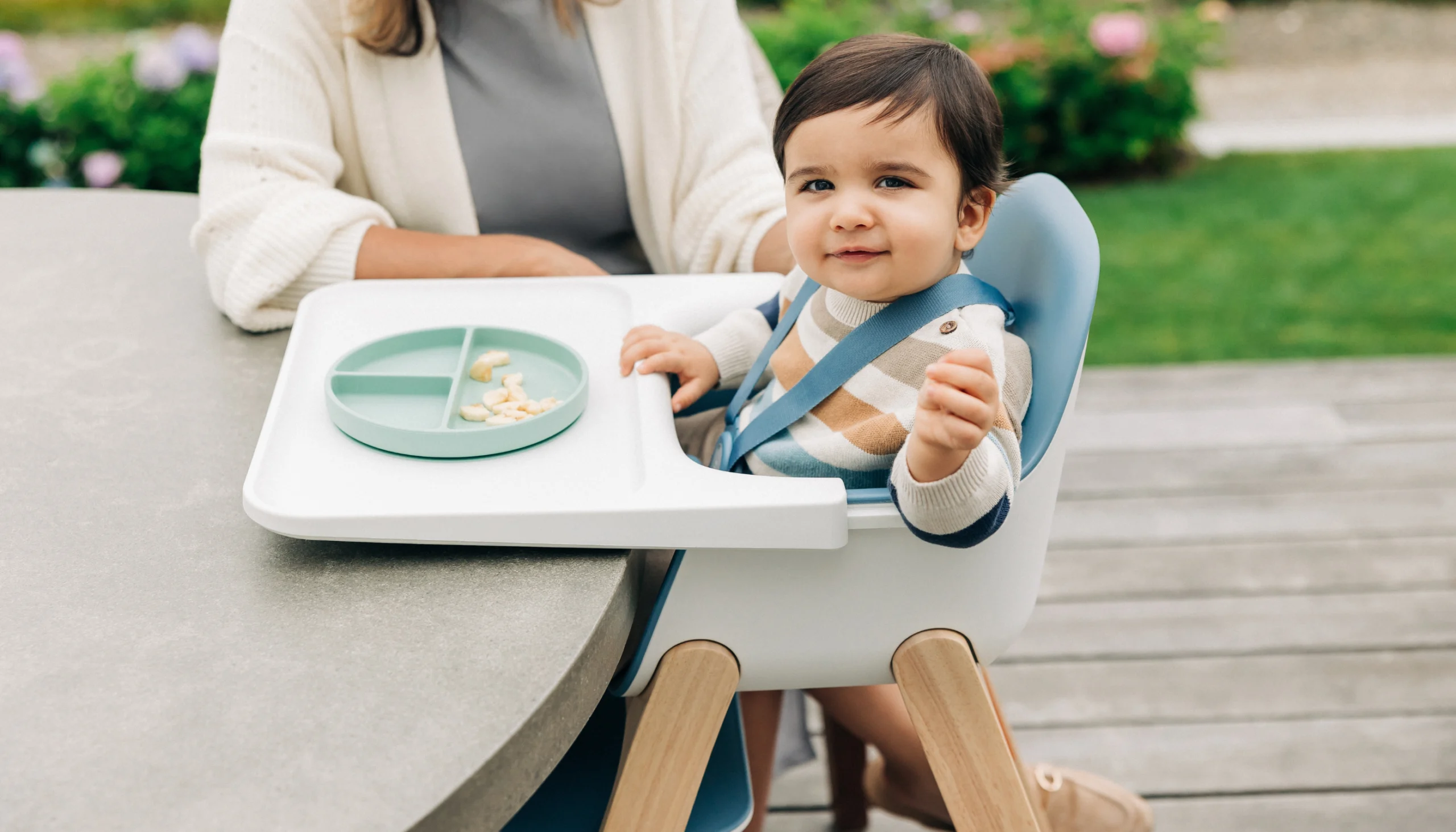 A child smiles in a Ciro High Chair, joining meal-time directly by sliding up and over a table, secured by the Ciro's easily adjustable harness