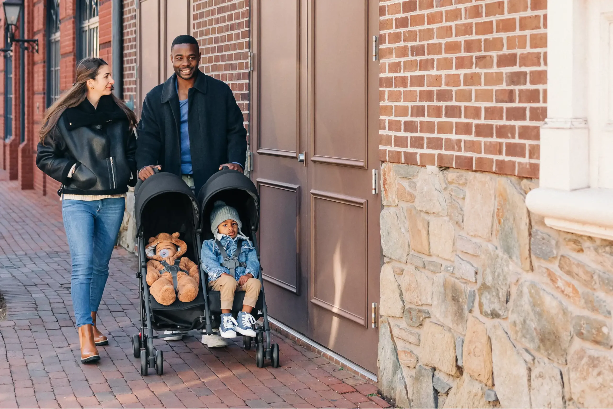 A family happily strolls with two children sitting side-by-side in the G-Link V2 relaxing at varied incline positions and riding comfortably due to the strollers all-aluminum frame and 4 sets of wheels (instead of six)