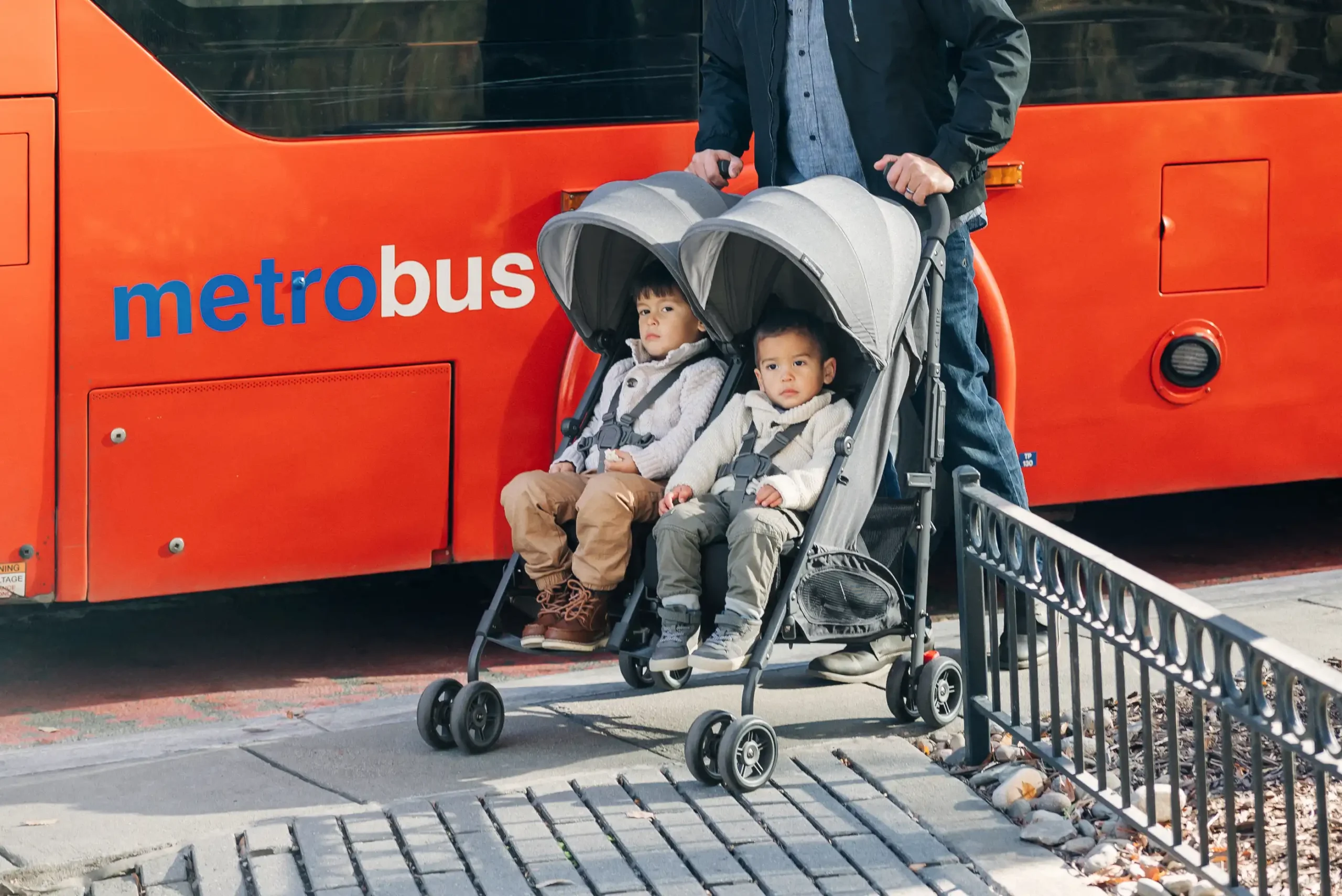 Two children happily stroll in a busy city in the G-Link V2, comfortably sitting side-by-side while the stroller is still able to fit through doorways!