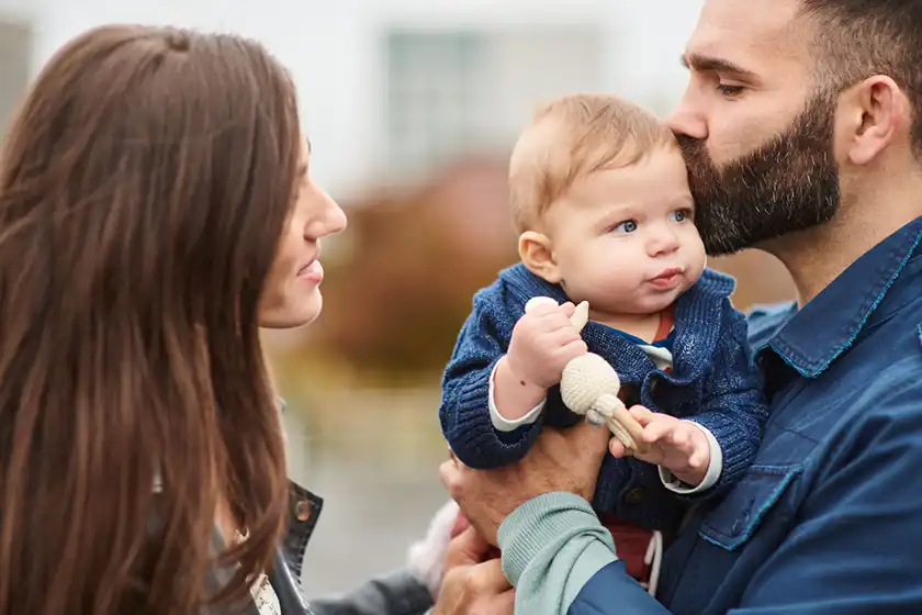 Mom and Dad stand close together while lovingly holding their infant - Mom smiling at Dad while he gently kisses the baby's head