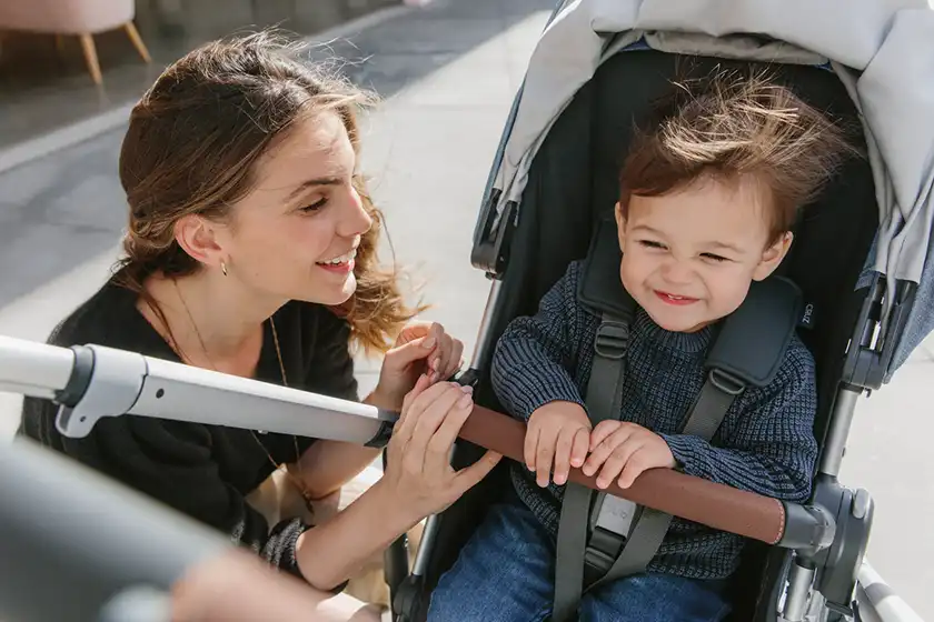 Smiling toddler sitting in Cruz Stroller, with hands on brown leather Bumper Bar. Mom leaning in and warmly smiles with child.