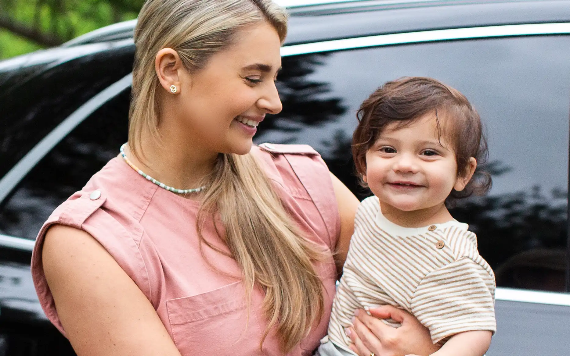 Parent standing while holding child outside of car, parent smiles and looks at smiling child