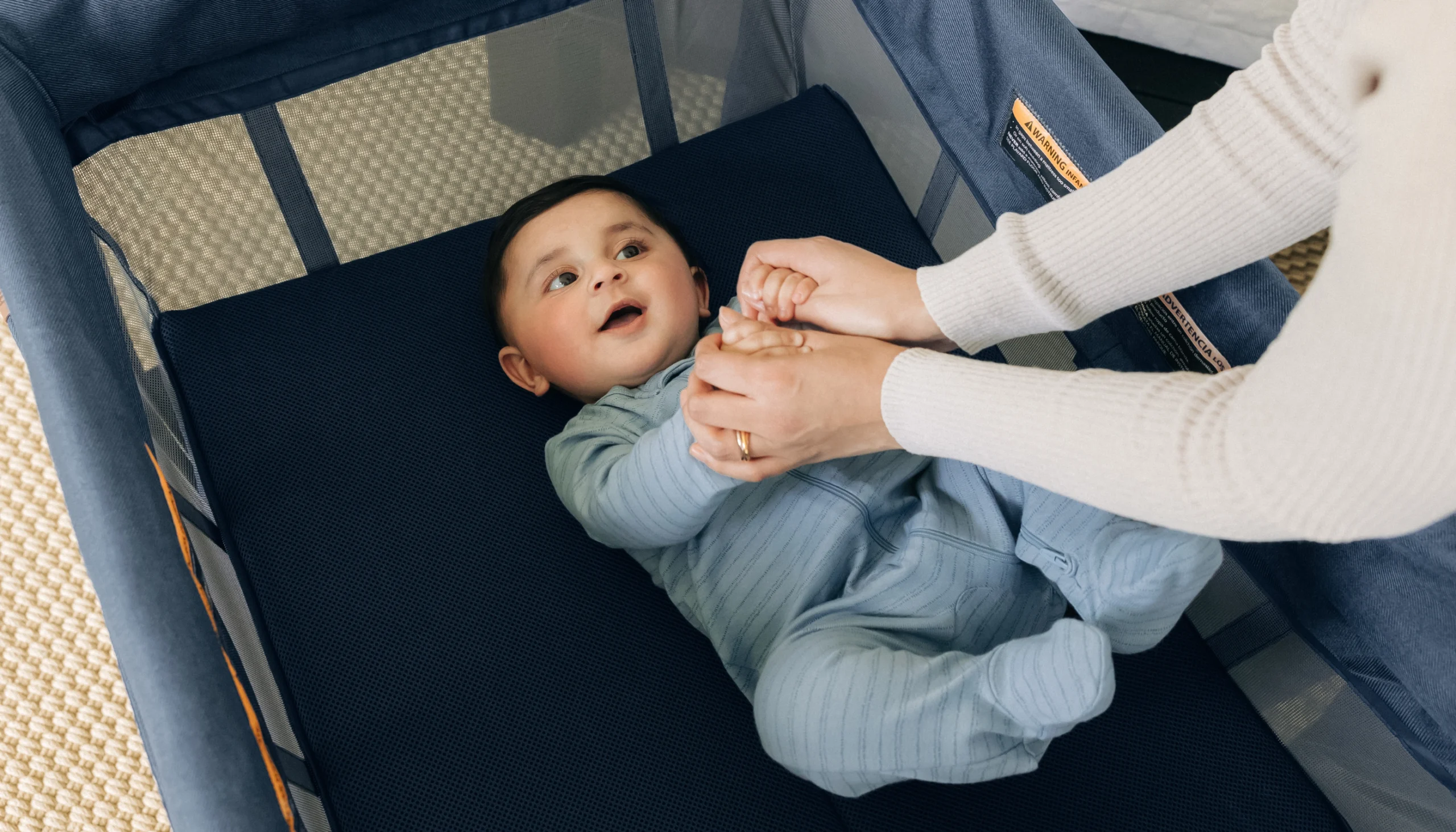 Baby holds his parent's hands playfully as he lays down comfortably in the Remi Playard